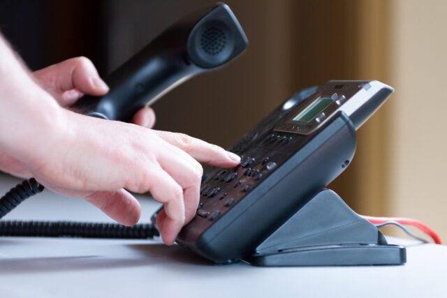Close-up of a person dialing a desk phone while holding the handset, with the keypad and display visible.