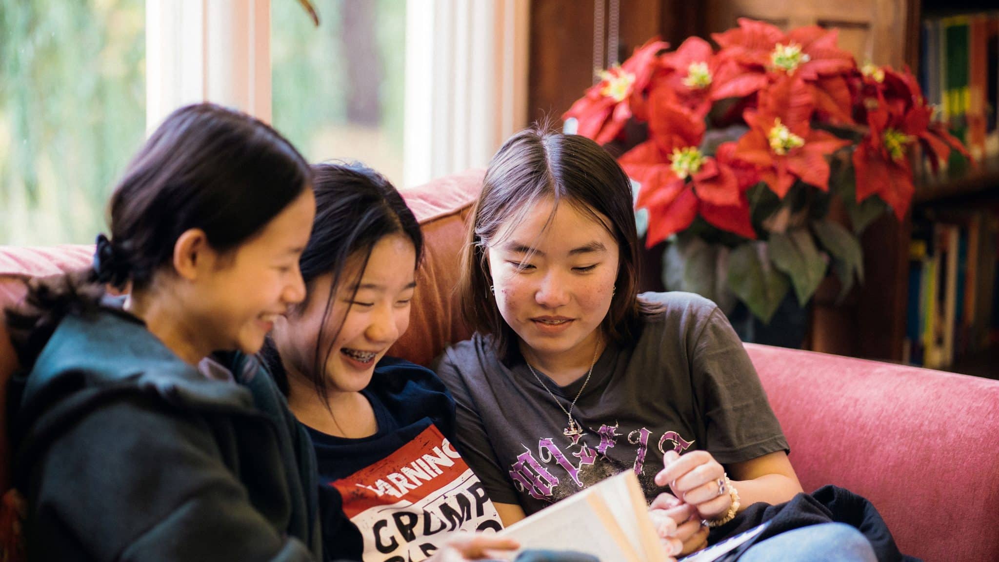 Three girls sit closely together on a sofa, smiling and looking at a book they are reading. Behind them is a window and a bright red poinsettia plant, creating a warm, cozy indoor setting.