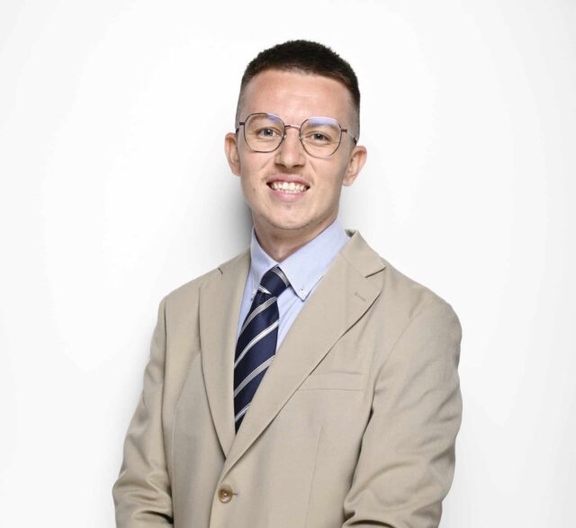 A young man wearing glasses stands against a light grey background, smiling at the camera. He is dressed in a beige blazer over a light blue shirt with a navy striped tie, and has his hands loosely clasped in front of him. He is also wearing a black smartwatch and dark trousers.