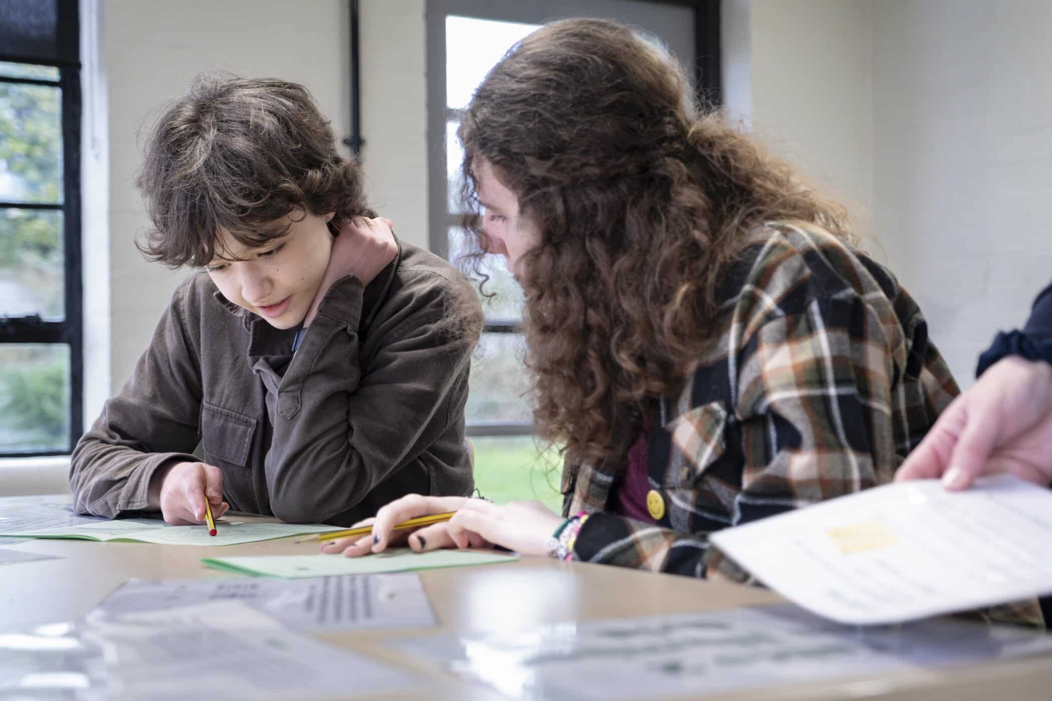 A young student and an older student or tutor sit side by side at a table indoors, concentrating on a worksheet together. The younger student holds a pencil and looks down at the paper while the older student leans in to help. Papers and notebooks are spread across the table, and daylight streams through large windows in the background.