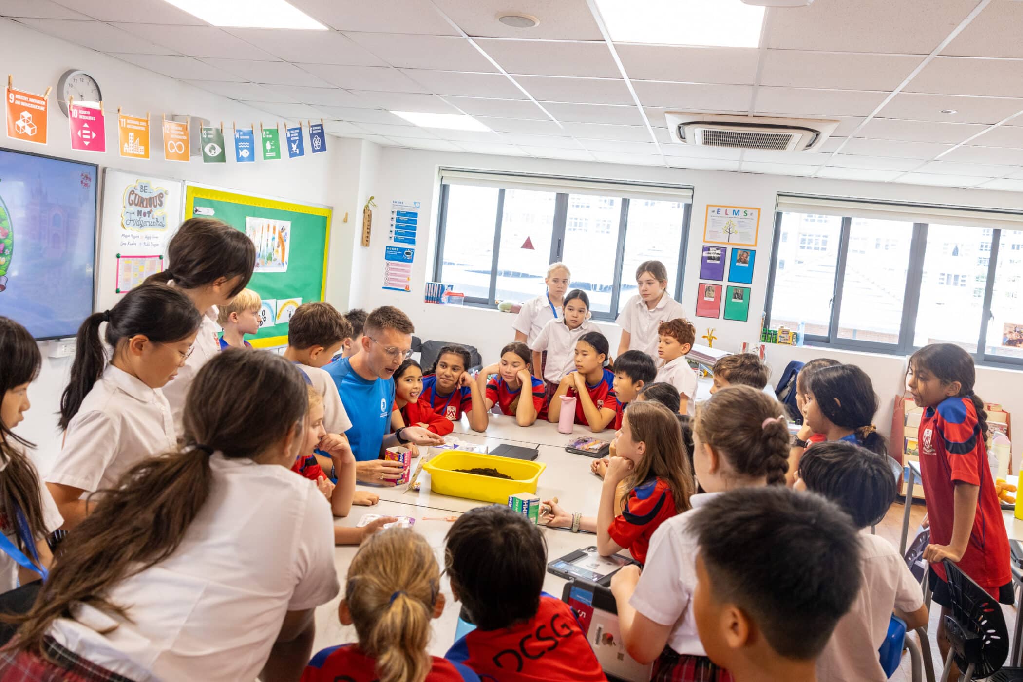 A teacher in a blue shirt leads a hands-on classroom activity with a large group of primary school children gathered around a table. The students, wearing a mix of white school uniforms and red-and-blue sports uniforms, watch attentively as the teacher demonstrates something using a yellow tray filled with soil. The bright classroom has large windows, educational posters, and colourful learning displays on the walls.