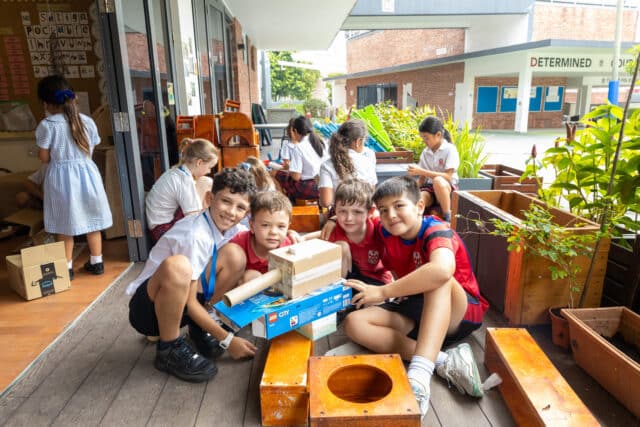 A group of primary school children sit together on a wooden outdoor learning deck, smiling at the camera while showing a creative model they have built from recycled materials, including cardboard boxes and tubes. Other students work and play in the background near classroom doors and planter boxes in a bright school courtyard.