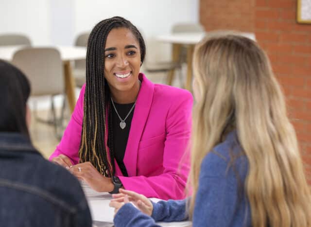 A staff member wearing a bright pink blazer sits at a table engaged in conversation with two students in a modern school setting. Smiling and attentive, she listens as the students speak, creating a welcoming and collaborative atmosphere. The background shows a light-filled communal learning space with tables and chairs.