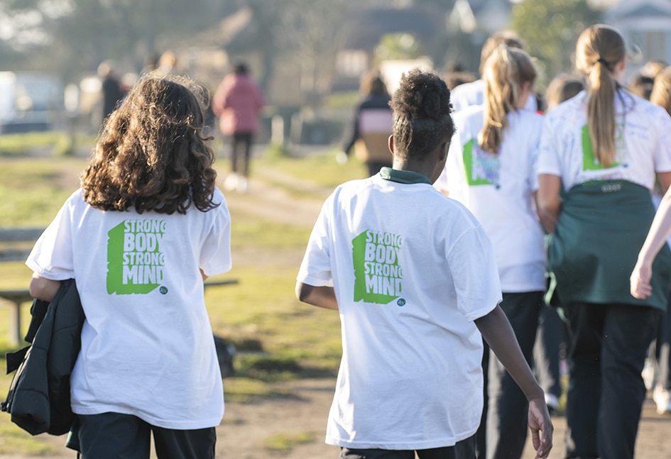 A group of school-aged children walk together along a sunlit path outdoors, viewed from behind. Several are wearing white T-shirts with the slogan “Strong Body Strong Mind” printed in green on the back, suggesting participation in a school wellbeing, fitness, or community event. The background is softly blurred, with more children and adults walking ahead.