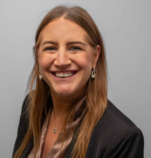 A smiling woman with long, straight, light brown hair, wearing a black blazer over a champagne-coloured blouse, silver earrings and a delicate necklace. She faces the camera against a plain grey background.