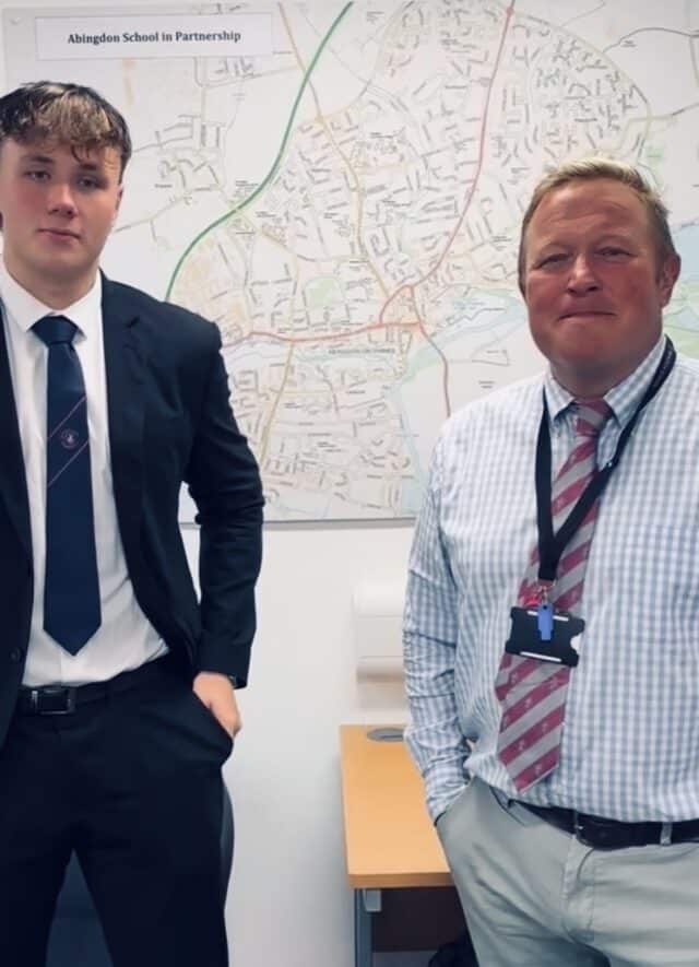 A student in a school blazer and tie stands beside a staff member wearing a checked shirt and striped tie in an office or classroom. Behind them is a large map on the wall with a sign reading “Abingdon School in Partnership.”