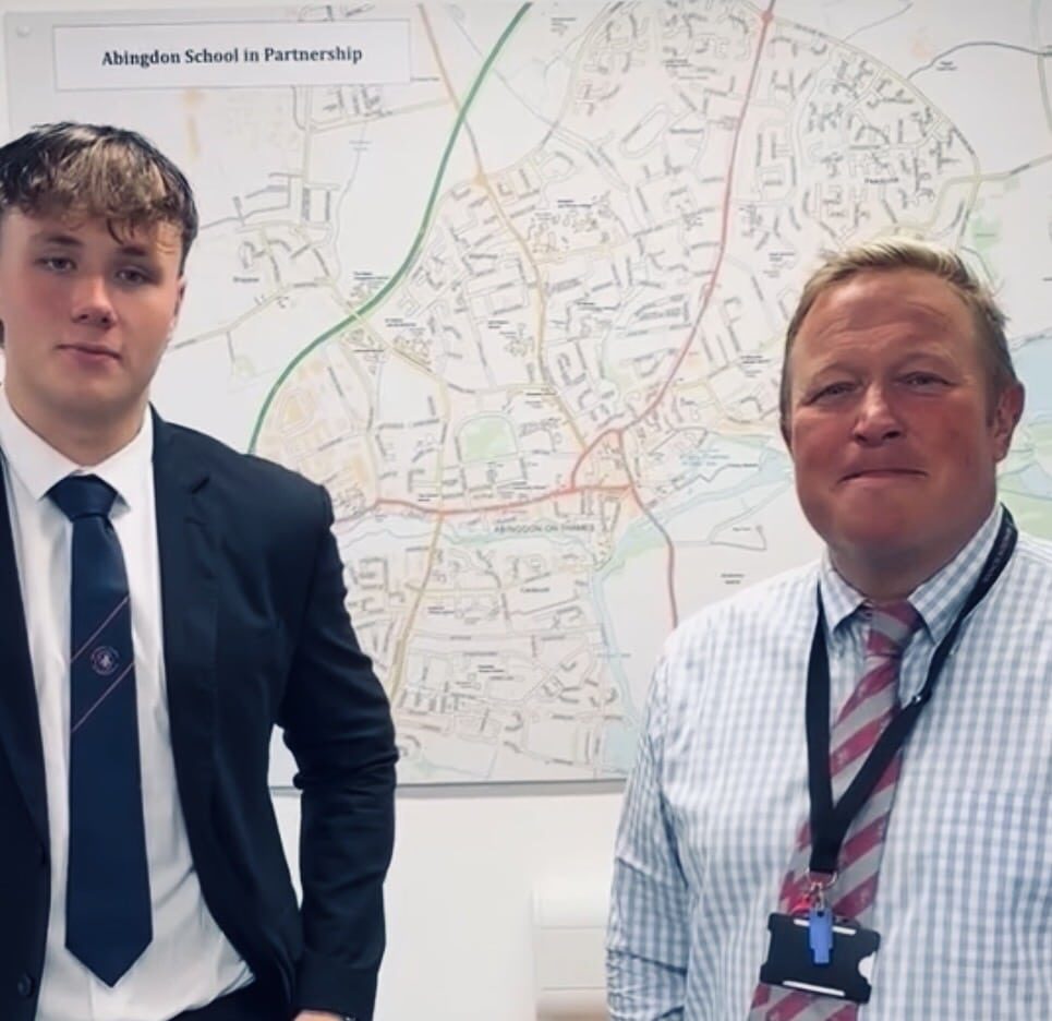 A student in a school blazer and tie stands beside a staff member wearing a checked shirt and striped tie in an office or classroom. Behind them is a large map on the wall with a sign reading “Abingdon School in Partnership.”