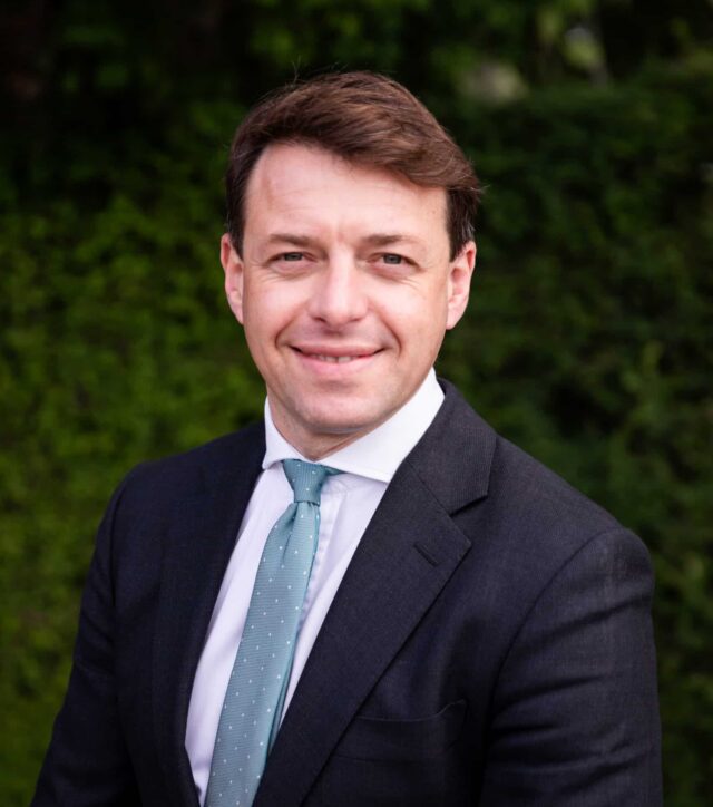 Professional head-and-shoulders portrait of a man wearing a dark suit, white shirt, and light blue tie. He is smiling slightly and facing the camera, with short brown hair neatly styled. The background is an out-of-focus wall of green foliage, creating a natural outdoor setting.