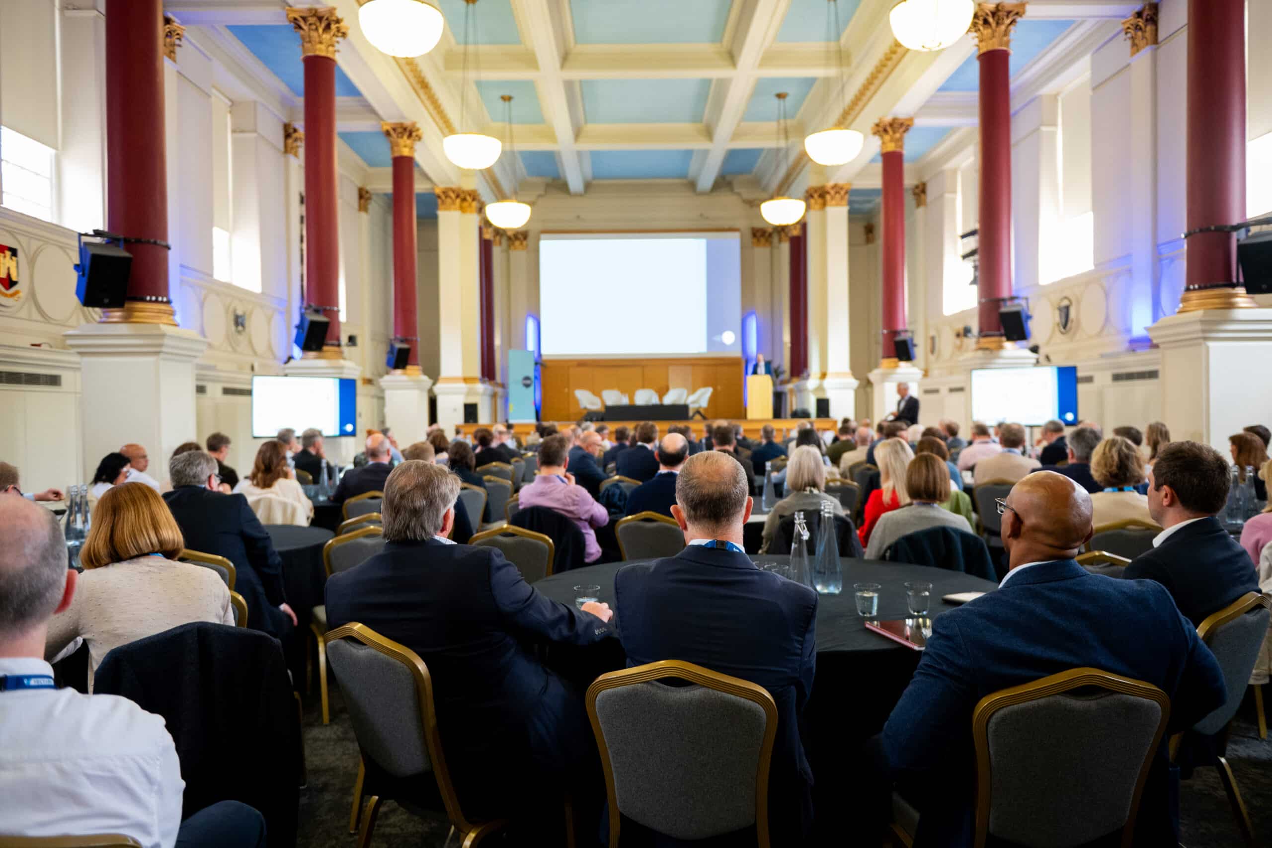 Audience seated at round tables in a large, elegant conference hall, facing a stage with a presentation screen and panel chairs. The room features high coffered ceilings painted light blue, tall red columns with gold capitals, and hanging globe lights. Attendees in business attire listen to a speaker at the front of the room during a professional conference or seminar.