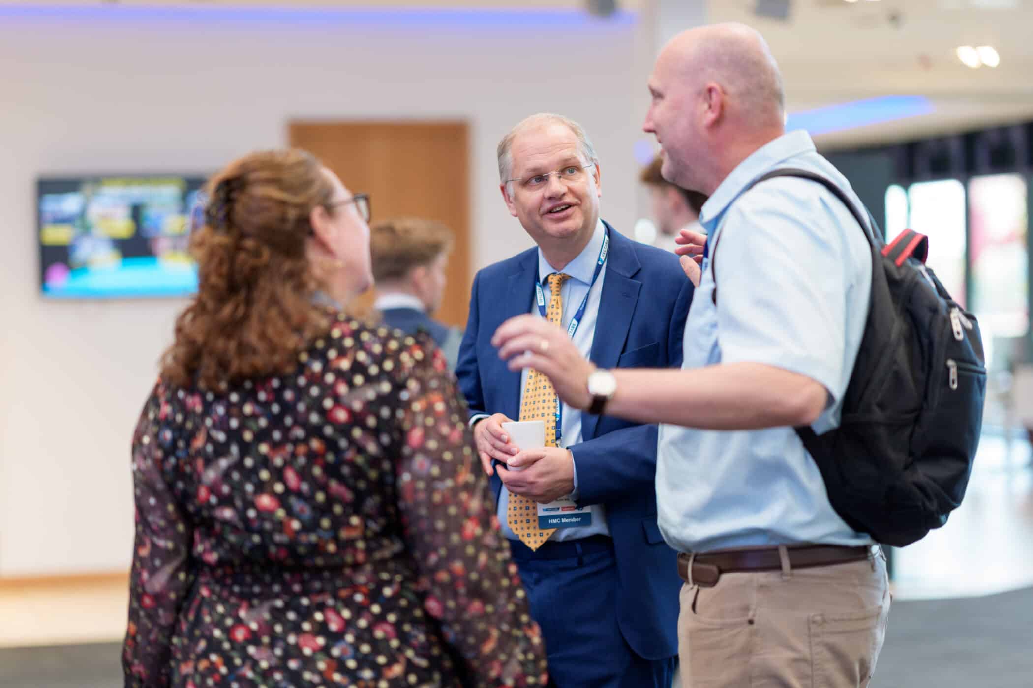 Three people chatting at a networking event, with a man in a blue suit speaking to a woman and another man wearing a backpack in a bright indoor conference setting.