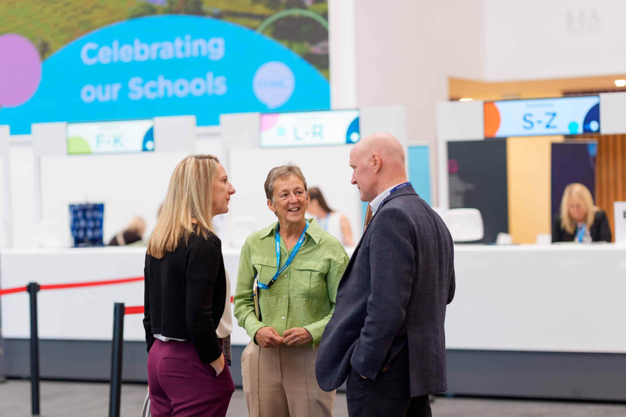 Three conference delegates stand in conversation in front of the registration desk at an HMC conference venue. Behind them, large signs reading 