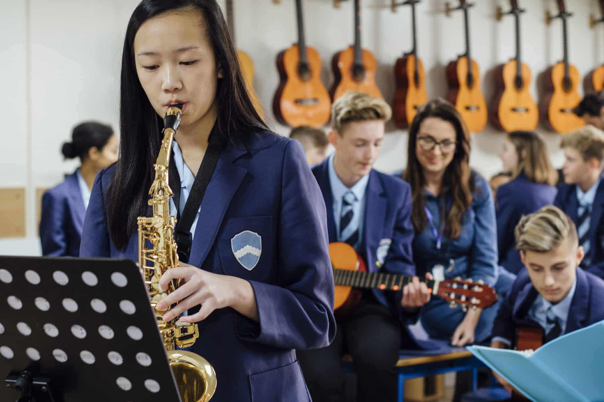 Student in school uniform playing a saxophone from sheet music during a music lesson, with a teacher and other students playing guitars in the background.