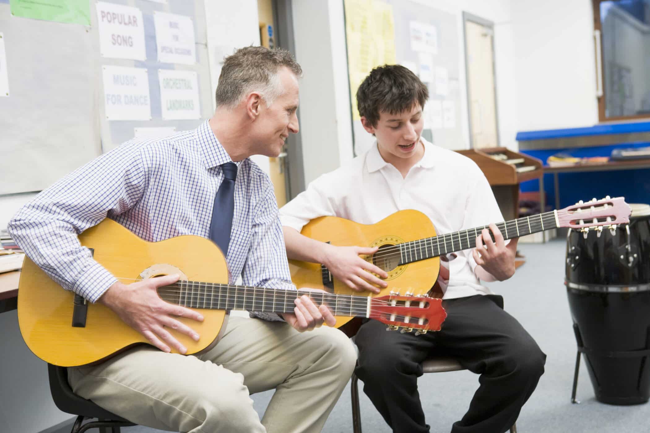 Music teacher and student sitting side by side playing acoustic guitars together during a classroom music lesson.