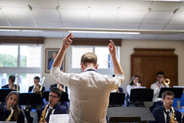Music teacher conducting a student ensemble, with pupils playing saxophones, trombones and other instruments in a bright music room.
