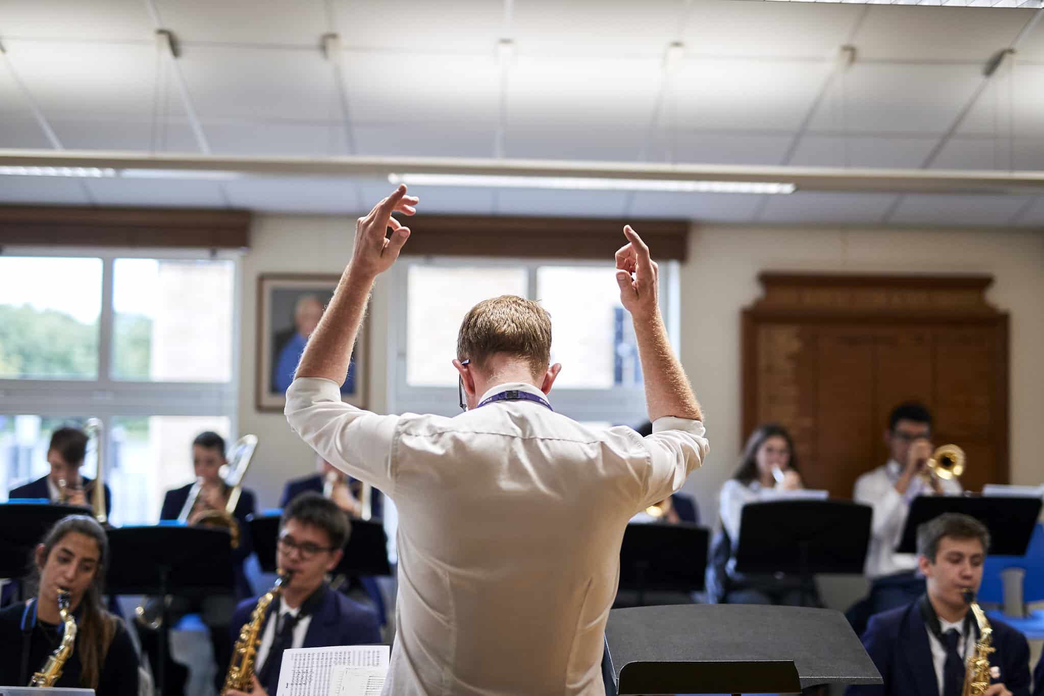 Music teacher conducting a student ensemble, with pupils playing saxophones, trombones and other instruments in a bright music room.