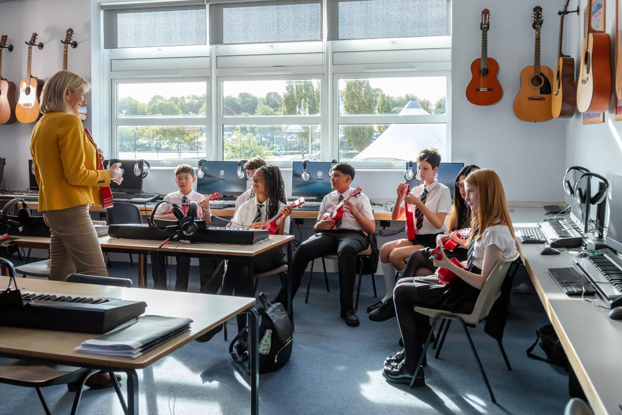 Music teacher leading a group of students playing ukuleles in a bright classroom equipped with keyboards, computers and guitars.