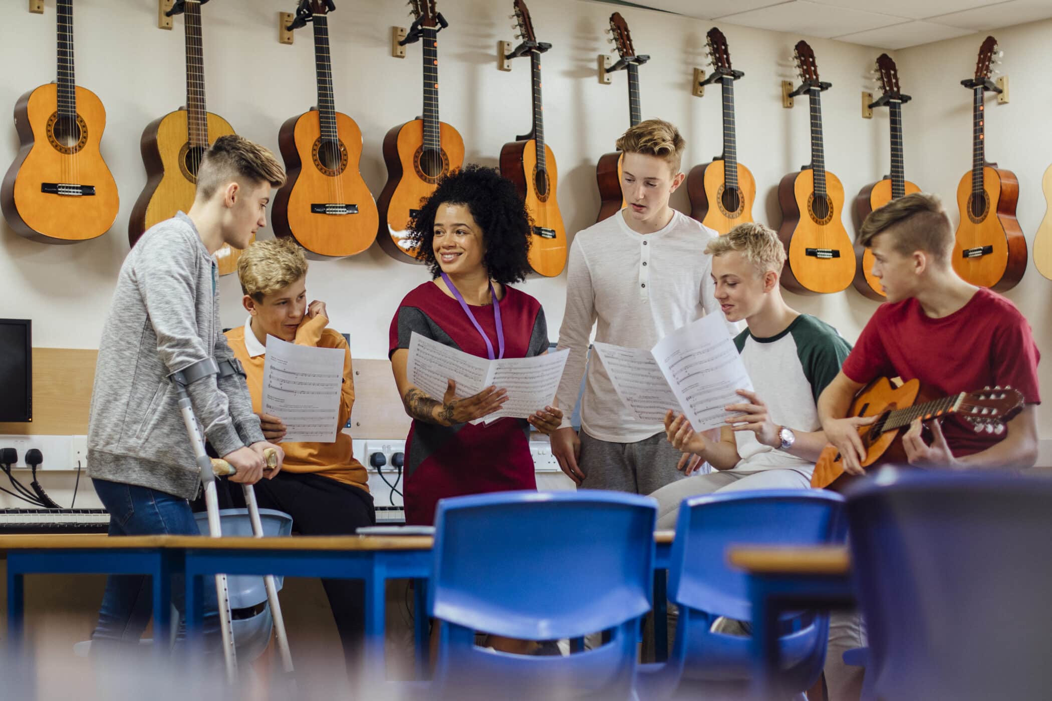Music teacher and five students gathered around sheet music in a classroom lined with guitars, with one student holding a guitar and another using crutches.