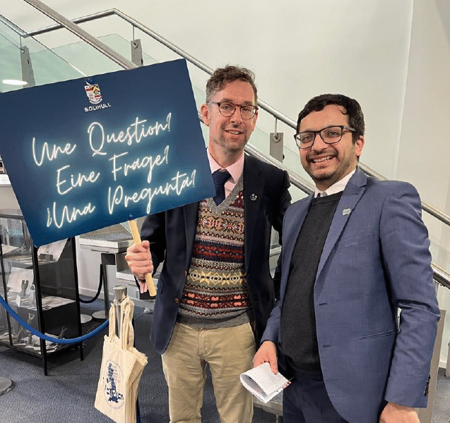 Charles Fillingham and a colleague at the National Festival of Languages at Solihull School. One holds a multilingual sign reading “Une Question! Eine Frage! ¡Una Pregunta!” while both smile at the camera.