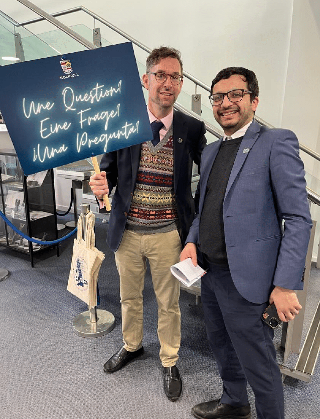 Charles Fillingham and a colleague at the National Festival of Languages at Solihull School. One holds a multilingual sign reading “Une Question! Eine Frage! ¡Una Pregunta!” while both smile at the camera.