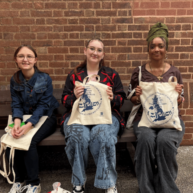 Three students sitting on a bench in front of a brick wall at the National Festival of Languages, with two holding cream festival tote bags printed with languages and place names.