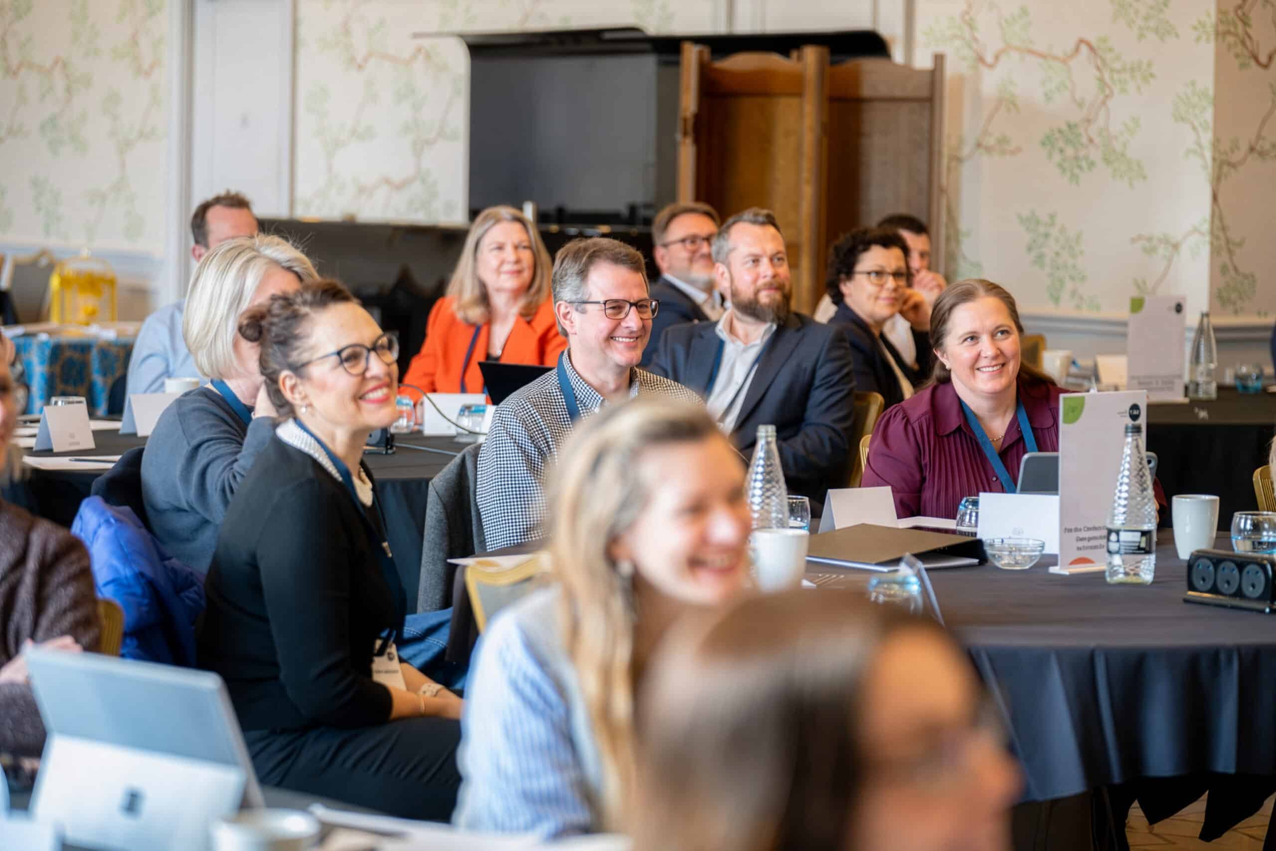 Conference delegates seated around round tables during a professional development session, smiling and listening attentively to a speaker off camera. Laptops, notebooks, name cards, water bottles and refreshments are visible on the tables, creating an engaged and collaborative learning environment.