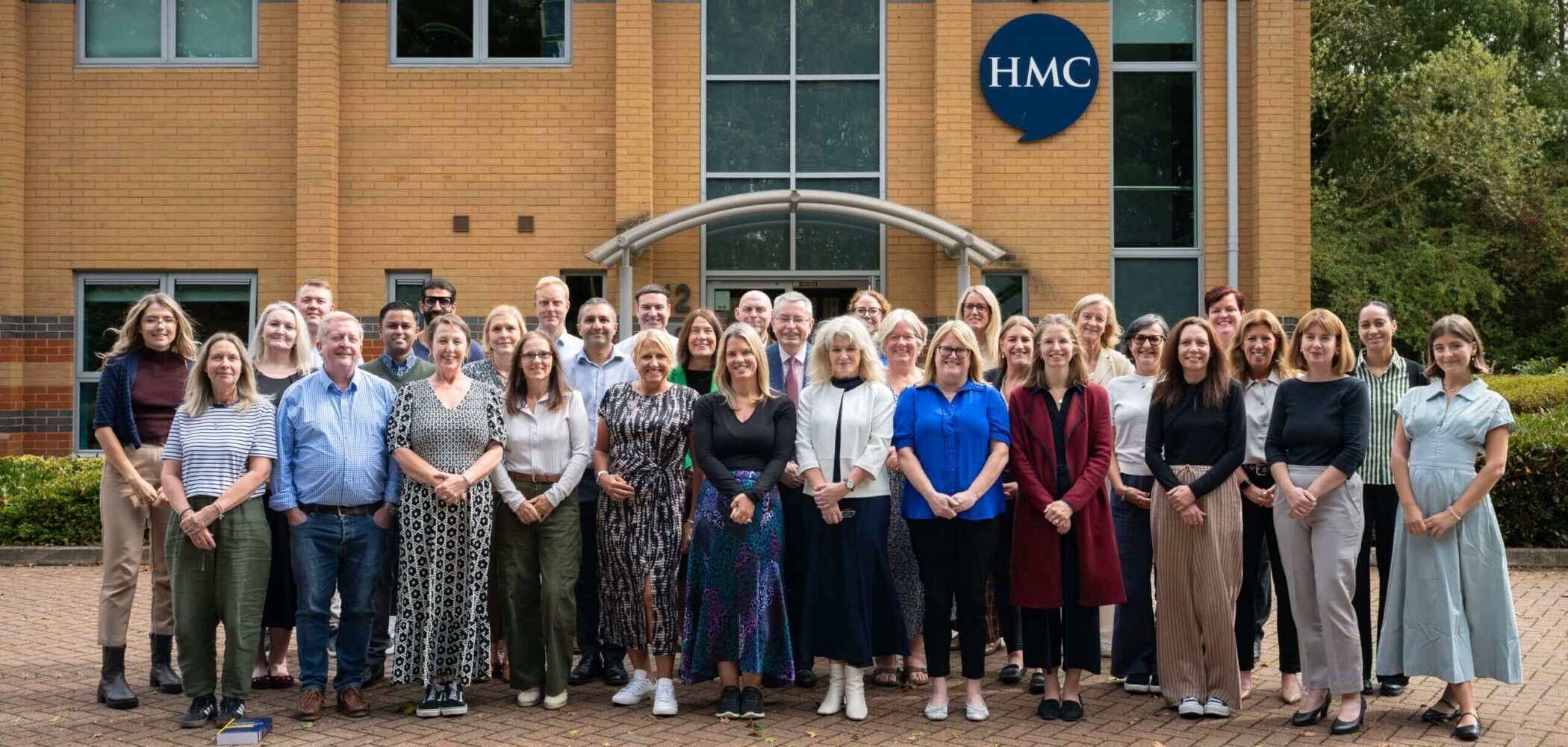 HMC staff group gathered outside the HMC office in Market Harborough, with the HMC logo visible on the building behind them.