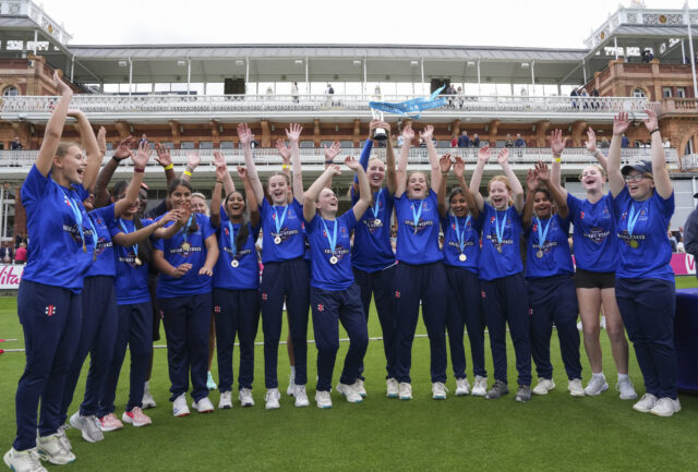 A girls' cricket team in blue kits celebrate with their trophy and medals at Lord's Cricket Ground following the Barclays Knight-Stokes Cup final.