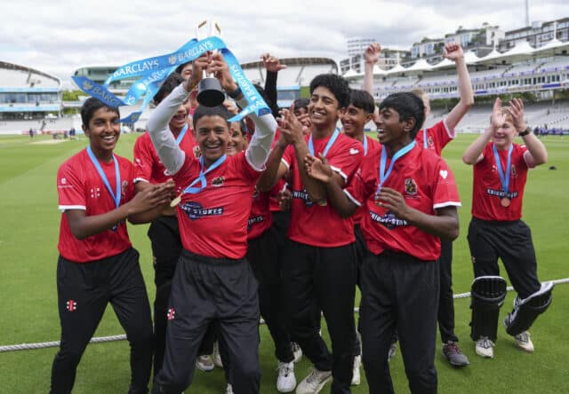 A boys' cricket team in red kits celebrate winning the Barclays Knight-Stokes Cup, raising their trophy at Lord's Cricket Ground.